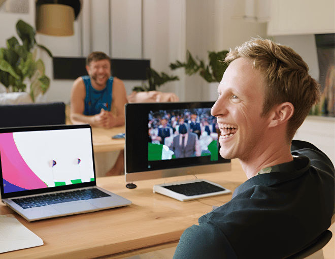 A white man, smiling, sits with a laptop open, celebrating energetically as he watches a thrilling cricket match unfold on a large television screen.